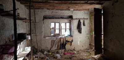 Rustic interior room showing stone and plaster walls, wooden ceiling beams, and various scattered items on the floor and shelves.