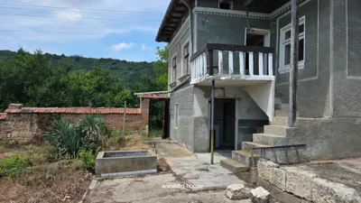 Two-storey house with stairs, balcony, brick and stone fence, concrete path, and plants in yard.