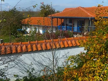 View of a white house with blue windows and a covered porch, surrounded by a flower garden, with a red tiled roof wall in the foreground.